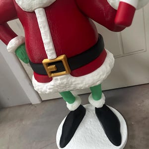 May include: A green character statue wearing a red coat with white trim, a black belt with a gold buckle, and black shoes. The statue has a white base and is holding a green and white object. The statue is a Christmas decoration.