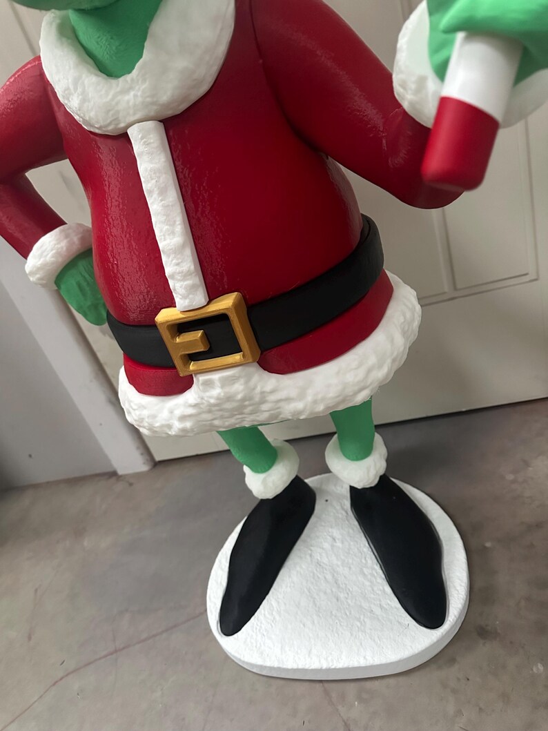 May include: A green character statue wearing a red coat with white trim, a black belt with a gold buckle, and black shoes. The statue has a white base and is holding a green and white object. The statue is a Christmas decoration.