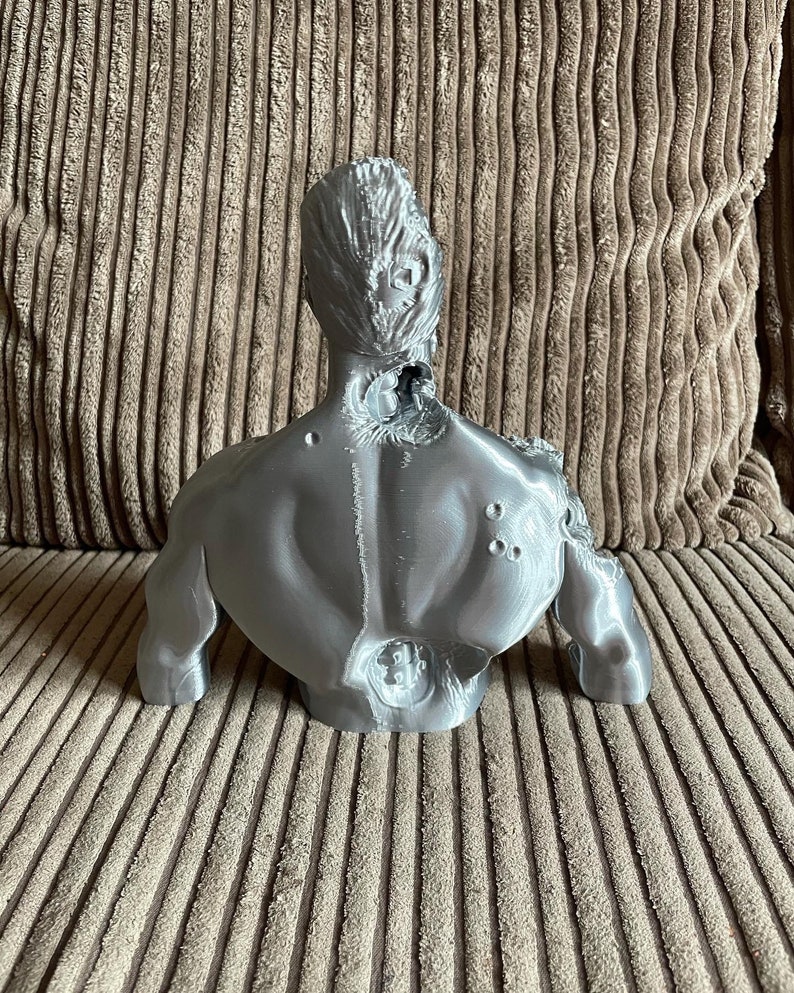 May include: A silver-colored 3D printed bust of a person with a damaged head and back. The bust is sitting on a brown textured surface.