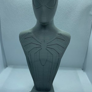 May include: A grey 3D printed bust of Spider-Man, wearing his red and blue suit with a black spider logo on the chest. The bust is on a base with the words "SPIDER-MAN" printed on it.