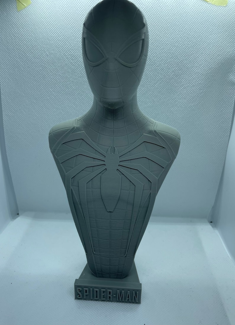 May include: A grey 3D printed bust of Spider-Man, wearing his red and blue suit with a black spider logo on the chest. The bust is on a base with the words "SPIDER-MAN" printed on it.