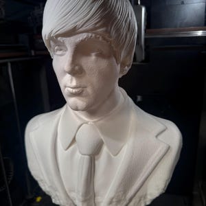 May include: A white bust sculpture of a man with a textured surface, wearing a suit jacket and tie. The sculpture has a detailed hairstyle and facial features. The bust is mounted on a blue base.