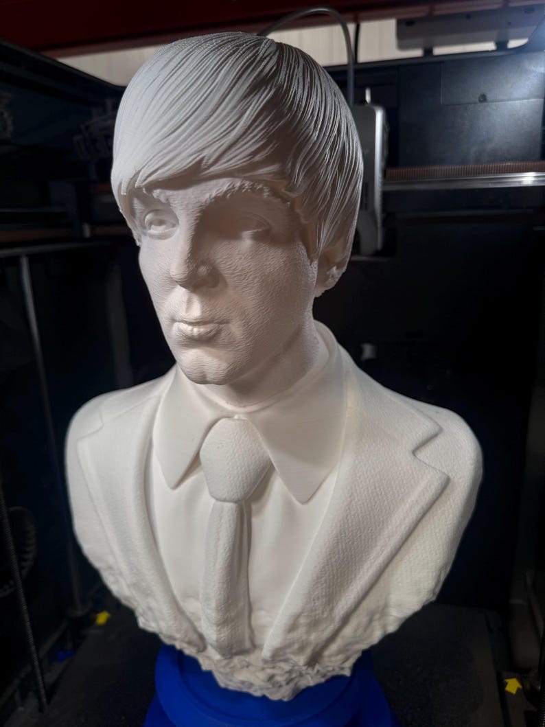 May include: A white bust sculpture of a man with a textured surface, wearing a suit jacket and tie. The sculpture has a detailed hairstyle and facial features. The bust is mounted on a blue base.