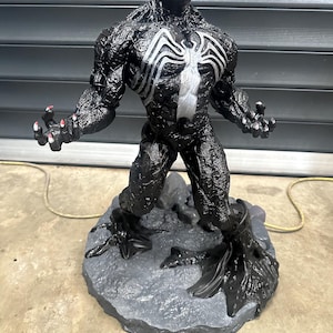 May include: A black Venom figurine with a white spider emblem on the chest. The figure has a detailed, textured appearance and stands on a gray, rocky base. The mouth is open, revealing red teeth and a red tongue.