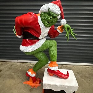 May include: A large, green figure of a character in a Santa Claus outfit, with a red coat, hat, and shoes. The figure is standing on a white and black box, with a mischievous expression. The figure is a Christmas decoration.