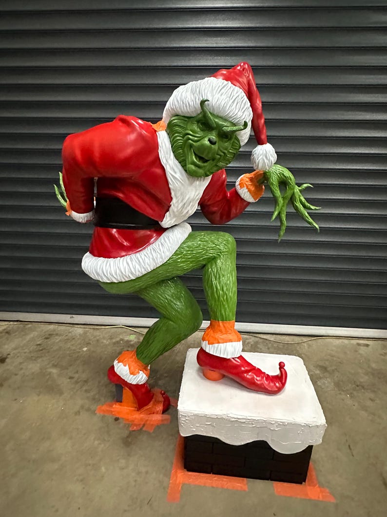 May include: A large, green figure of a character in a Santa Claus outfit, with a red coat, hat, and shoes. The figure is standing on a white and black box, with a mischievous expression. The figure is a Christmas decoration.
