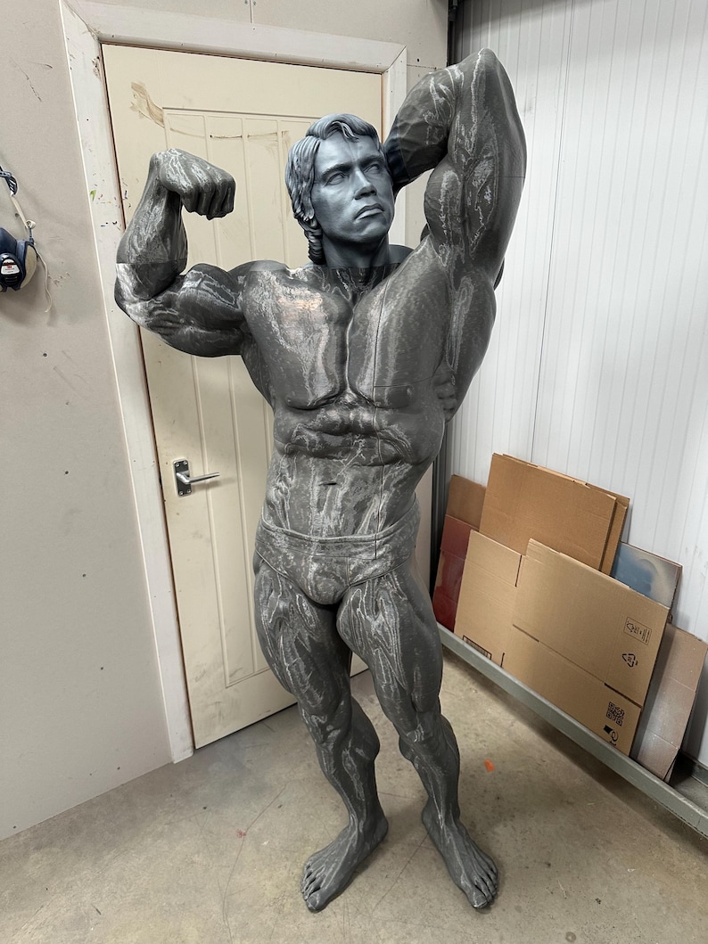 May include: A gray statue of a bodybuilder flexing his muscles. The statue is a detailed representation of a muscular man, with defined abs and arms. The statue is standing in a room with a white door and cardboard boxes in the background.