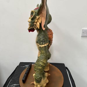 May include: A detailed side view of a Gremlin statue, featuring a green and tan color scheme. The statue has large ears, a textured body, and a menacing expression. It stands on a brown circular base with a black rectangular piece.