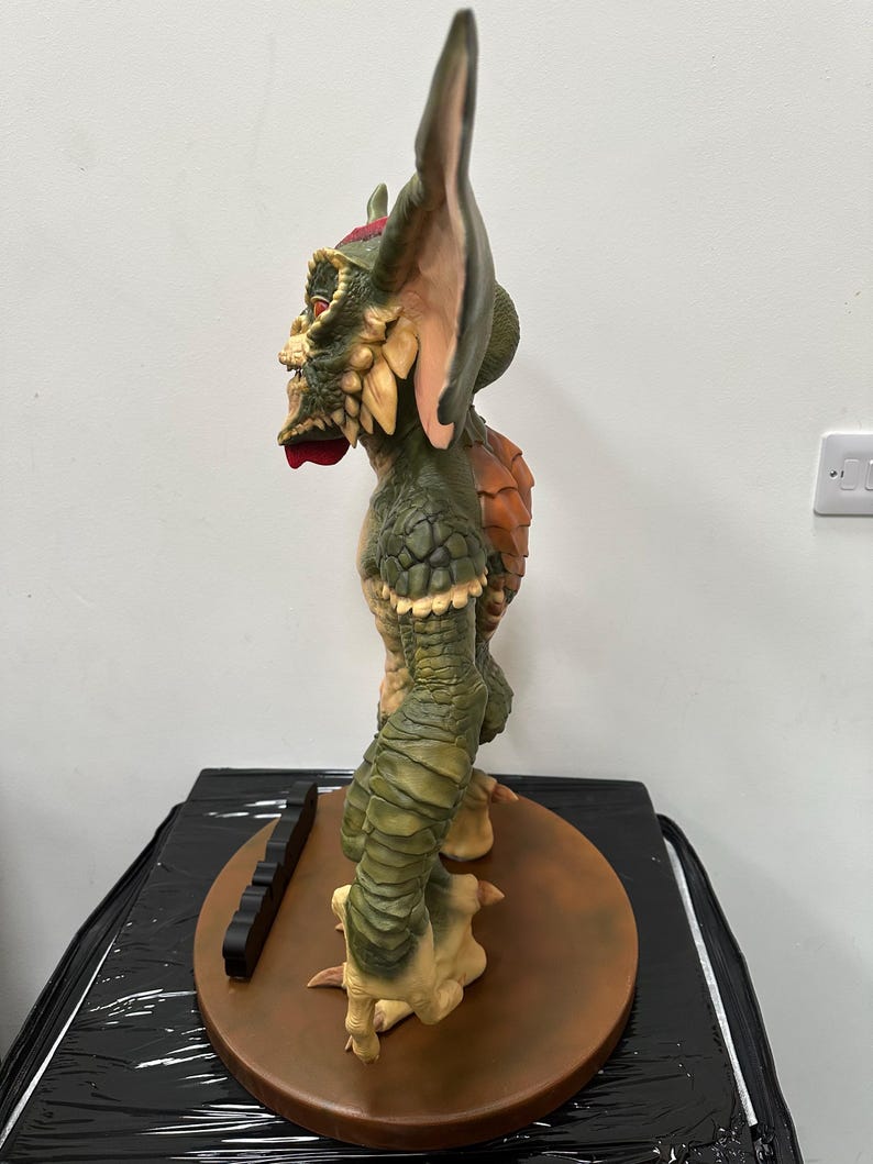 May include: A detailed side view of a Gremlin statue, featuring a green and tan color scheme. The statue has large ears, a textured body, and a menacing expression. It stands on a brown circular base with a black rectangular piece.