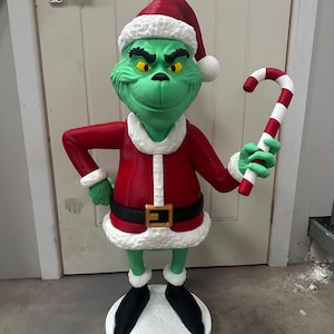 May include: A green Grinch figure wearing a red Santa suit with white trim and a black belt with a gold buckle. The Grinch is holding a red and white candy cane. The figure is standing on a white base and wearing a Santa hat.