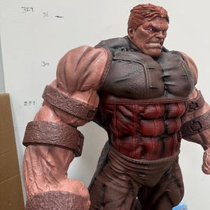 May include: A detailed statue of a muscular, red-skinned character with a stern expression. The figure has brown hair and is wearing a brown and red outfit with large, textured armbands. The statue is approximately 2 feet tall.