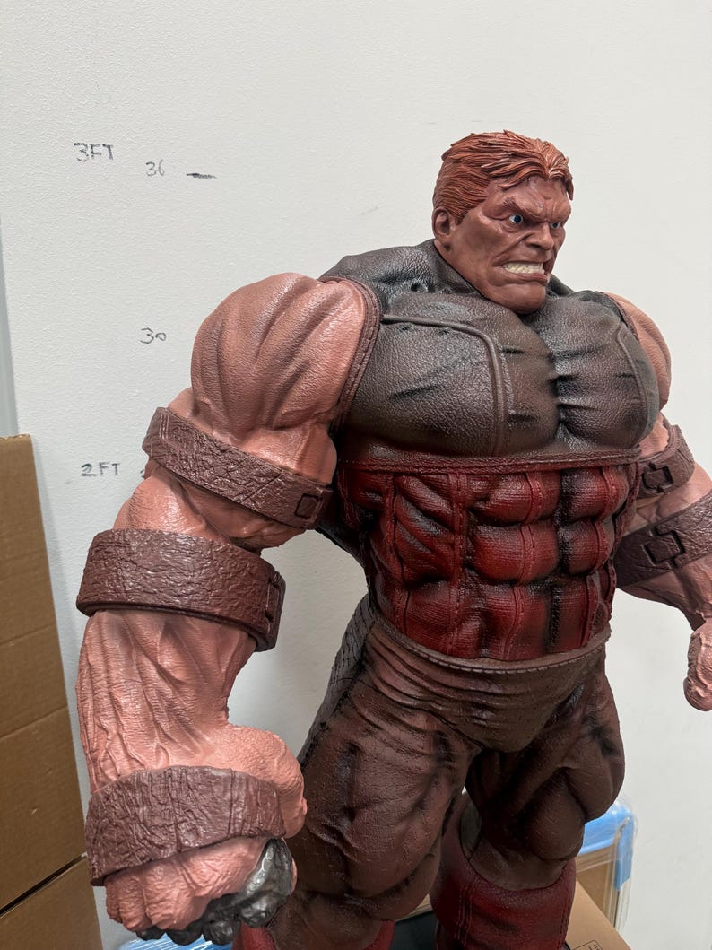 May include: A detailed statue of a muscular, red-skinned character with a stern expression. The figure has brown hair and is wearing a brown and red outfit with large, textured armbands. The statue is approximately 2 feet tall.
