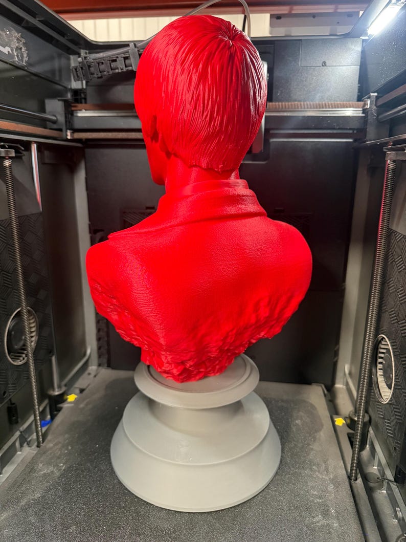 May include: A bright red bust of a person, viewed from the back, with short hair and a collared shirt. The bust is mounted on a gray, tiered pedestal. The sculpture is inside a 3D printer.