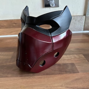 May include: A burgundy and black plastic mask with two holes on the side. The mask has a raised design on the front.