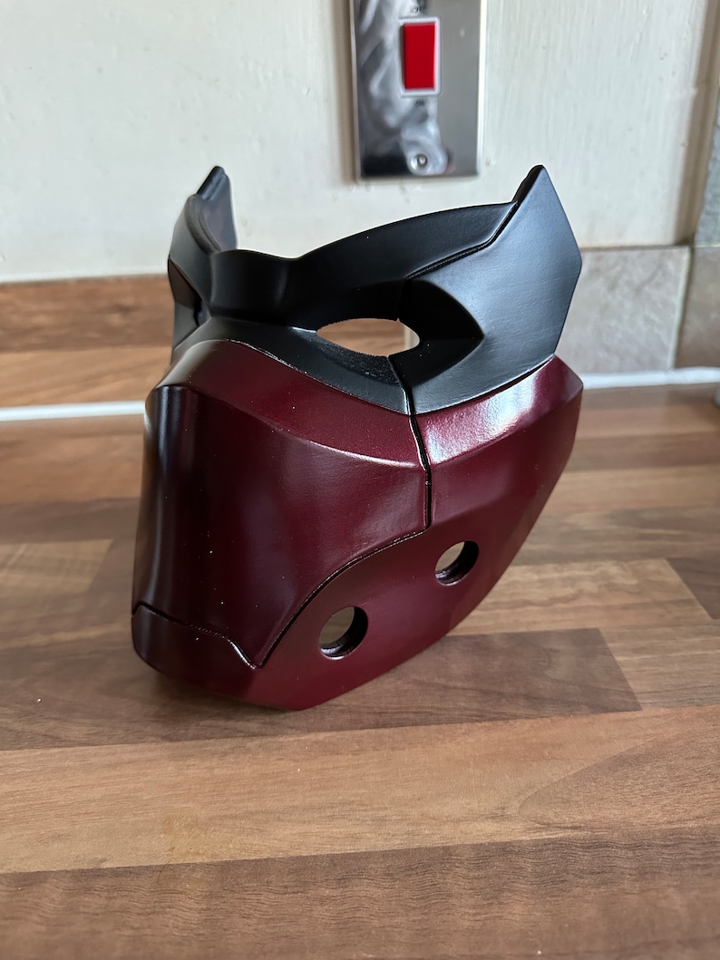 May include: A burgundy and black plastic mask with two holes on the side. The mask has a raised design on the front.
