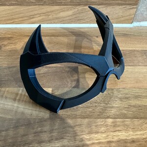 Puede incluir: Una m&aacute;scara de pl&aacute;stico negro con un dise&ntilde;o &uacute;nico, que se asemeja a una forma de ojo estilizada.
