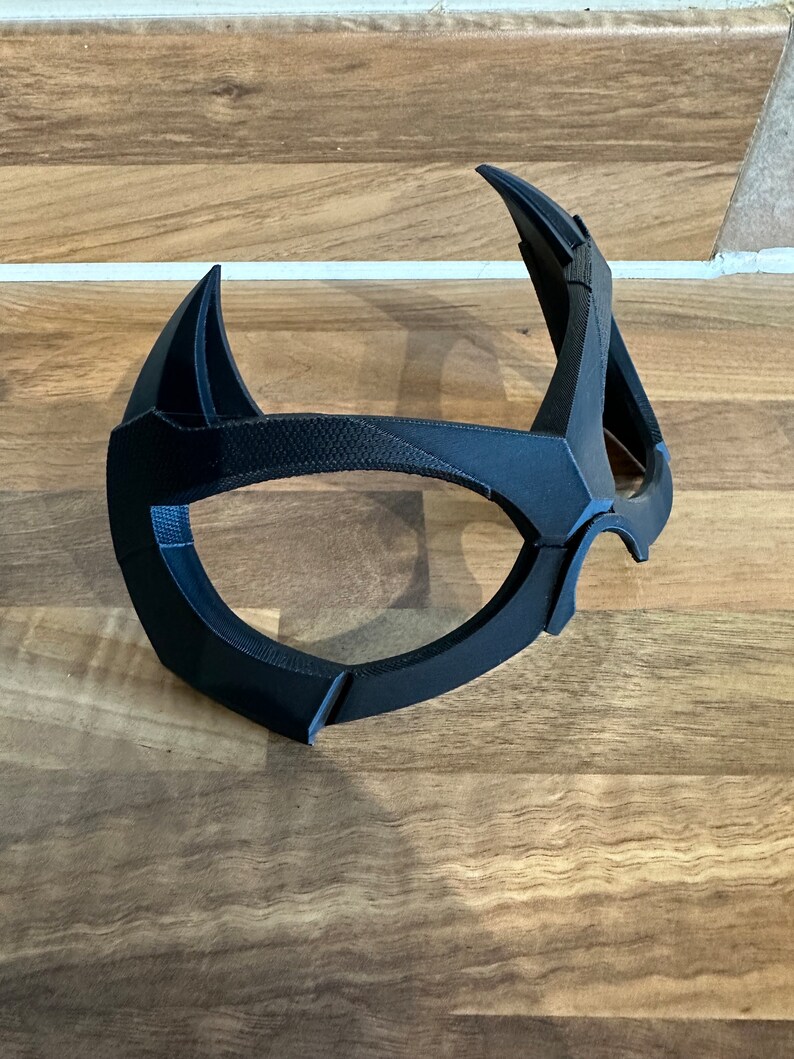 Puede incluir: Una m&aacute;scara de pl&aacute;stico negro con un dise&ntilde;o &uacute;nico, que se asemeja a una forma de ojo estilizada.