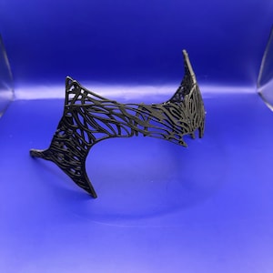 May include: A black, decorative crown with an intricate, openwork pattern. The crown has pointed arches and a flowing, organic design. The crown is made of a dark material and is set against a blue background.