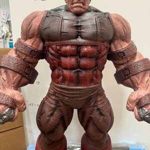May include: A detailed figurine of a muscular character, predominantly red and brown. The figure has a stern expression, with a detailed face and hair. The costume includes a chest piece, pants, and arm bands. The figure is standing upright.