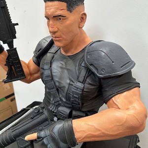 May include: A detailed bust of a muscular figure with short, dark hair and a stern expression. The figure wears a black tactical shirt, shoulder armor, and fingerless gloves, and holds a black rifle. The bust is painted in realistic tones.