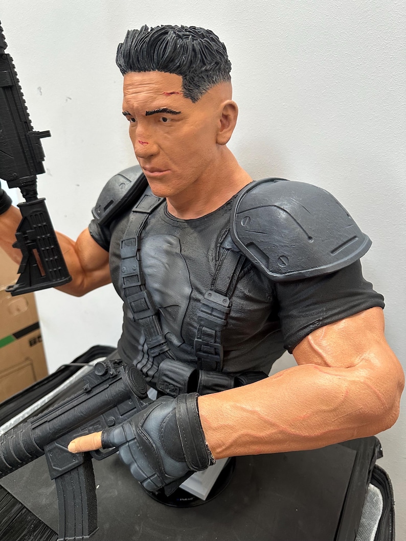 May include: A detailed bust of a muscular figure with short, dark hair and a stern expression. The figure wears a black tactical shirt, shoulder armor, and fingerless gloves, and holds a black rifle. The bust is painted in realistic tones.