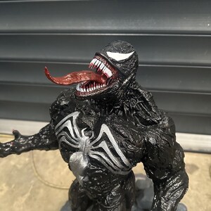 May include: A detailed figurine of the Marvel Comics character Venom. The figure is predominantly black with white accents, including the iconic spider-symbol on the chest and the eyes. The mouth is open, revealing sharp white teeth and a long, red tongue.