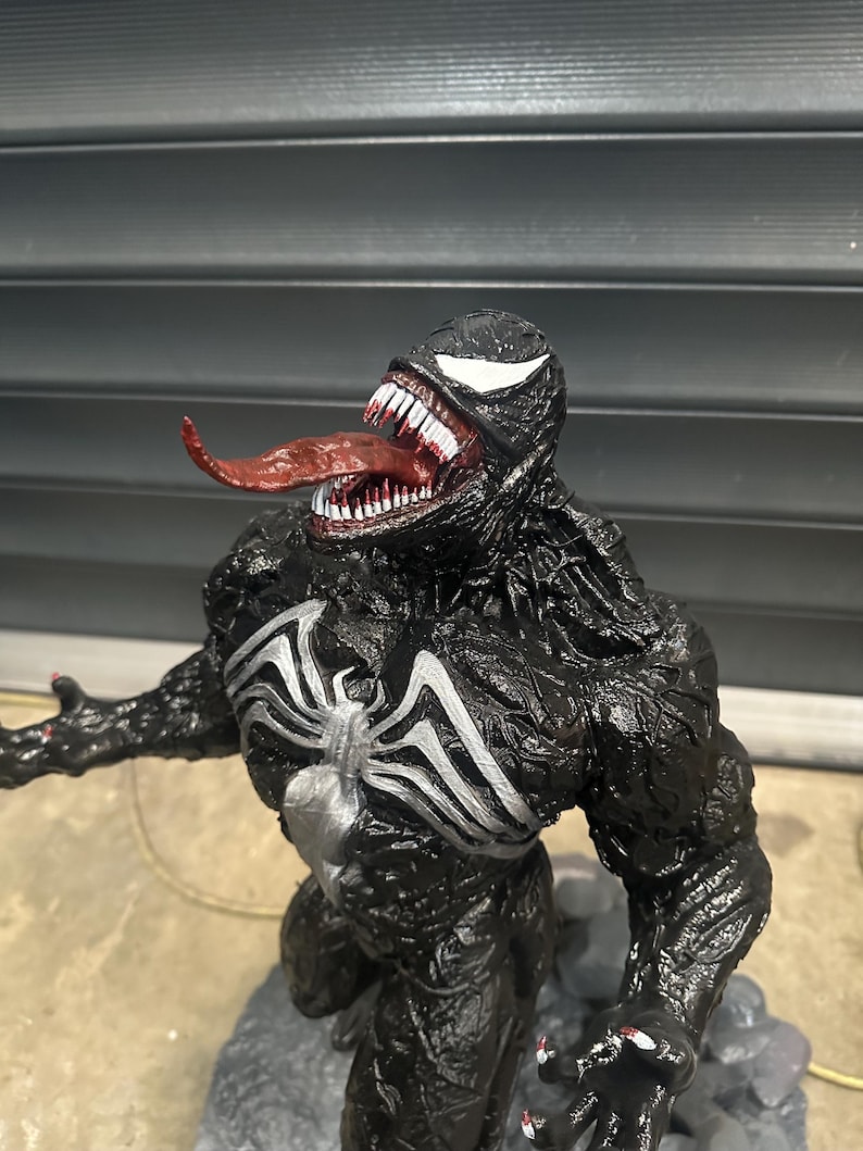 May include: A detailed figurine of the Marvel Comics character Venom. The figure is predominantly black with white accents, including the iconic spider-symbol on the chest and the eyes. The mouth is open, revealing sharp white teeth and a long, red tongue.