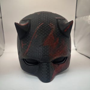 May include: A black and red helmet with two pointed horns. The helmet has a textured surface and eye coverings. Red accents are visible on the horns and the lower portion of the helmet.