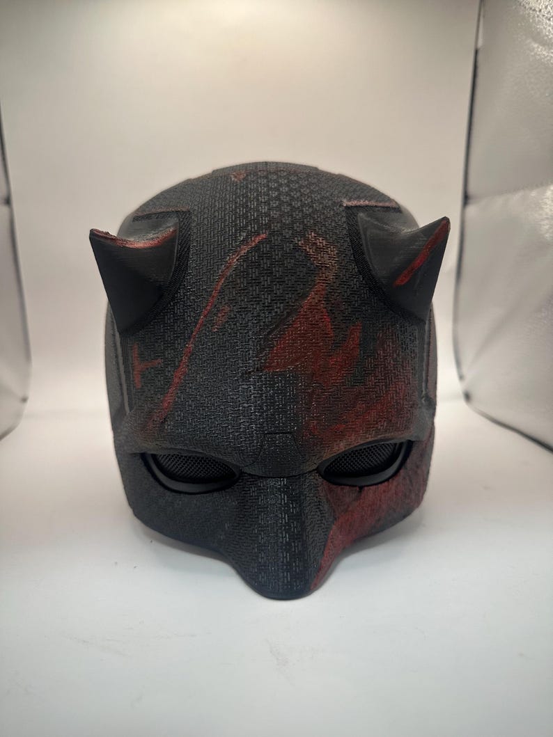 May include: A black and red helmet with two pointed horns. The helmet has a textured surface and eye coverings. Red accents are visible on the horns and the lower portion of the helmet.