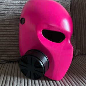 K&ouml;nnte beinhalten: Eine rosafarbene Gasmaske mit einem schwarzen Filter. Die Maske hat ein gro&szlig;es Augenloch und ein kleines Loch an der Seite.