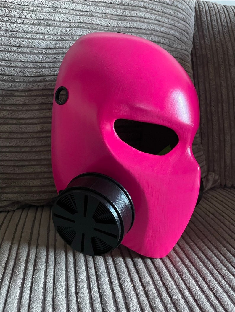 K&ouml;nnte beinhalten: Eine rosafarbene Gasmaske mit einem schwarzen Filter. Die Maske hat ein gro&szlig;es Augenloch und ein kleines Loch an der Seite.