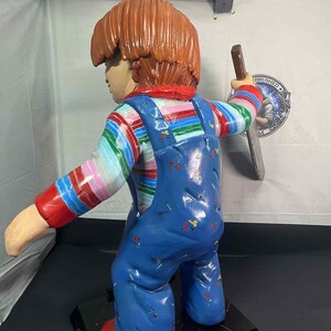 May include: A large, painted figurine of a character with orange hair, wearing a striped shirt, blue overalls, and red shoes. The figure is holding a knife. The base is black with red accents.