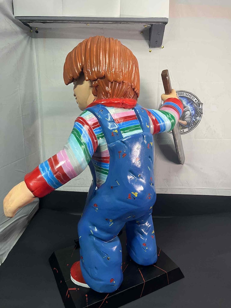May include: A large, painted figurine of a character with orange hair, wearing a striped shirt, blue overalls, and red shoes. The figure is holding a knife. The base is black with red accents.