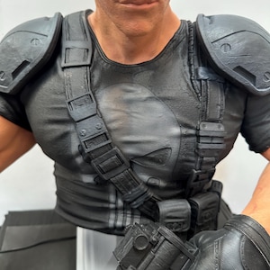 May include: A detailed bust of a character in a black t-shirt with a white skull emblem. The figure wears tactical gear, including shoulder pads, straps, and gloves. The figure is holding a black rifle.