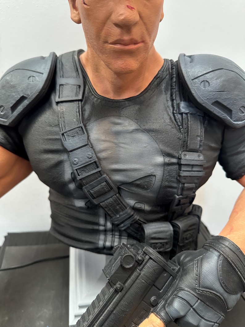 May include: A detailed bust of a character in a black t-shirt with a white skull emblem. The figure wears tactical gear, including shoulder pads, straps, and gloves. The figure is holding a black rifle.