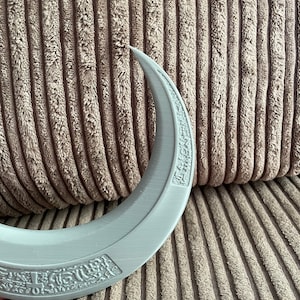 May include: A gray crescent moon-shaped object with a raised design and text around the edge. The object is made of a smooth, plastic-like material.
