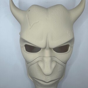 May include: A light beige, horned mask with an angry expression. The mask has large eye openings, a prominent nose, and a defined chin. The horns curve upwards and outwards. The mask appears to be made of a textured material.