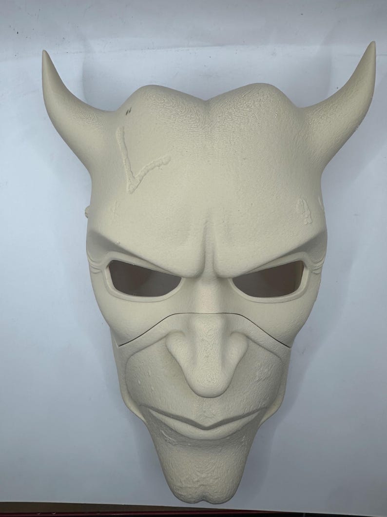 May include: A light beige, horned mask with an angry expression. The mask has large eye openings, a prominent nose, and a defined chin. The horns curve upwards and outwards. The mask appears to be made of a textured material.