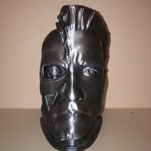 May include: A 3D printed bust of a robot with a human face. The robot's face is partially exposed, revealing mechanical parts. The bust is painted in a metallic silver color.