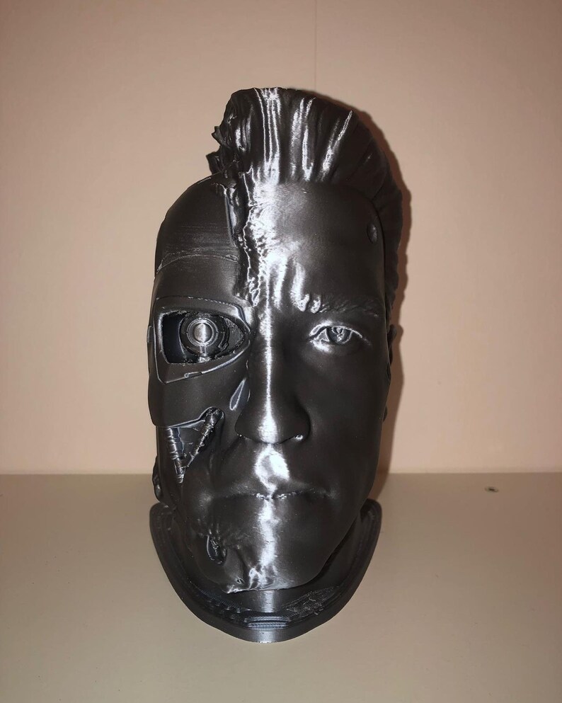 May include: A 3D printed bust of a robot with a human face. The robot's face is partially exposed, revealing mechanical parts. The bust is painted in a metallic silver color.