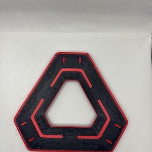 May include: A black and red geometric object with a triangular shape and a textured surface. The object has a red border and red accents within the black interior. The design features a series of parallel lines and rounded shapes.