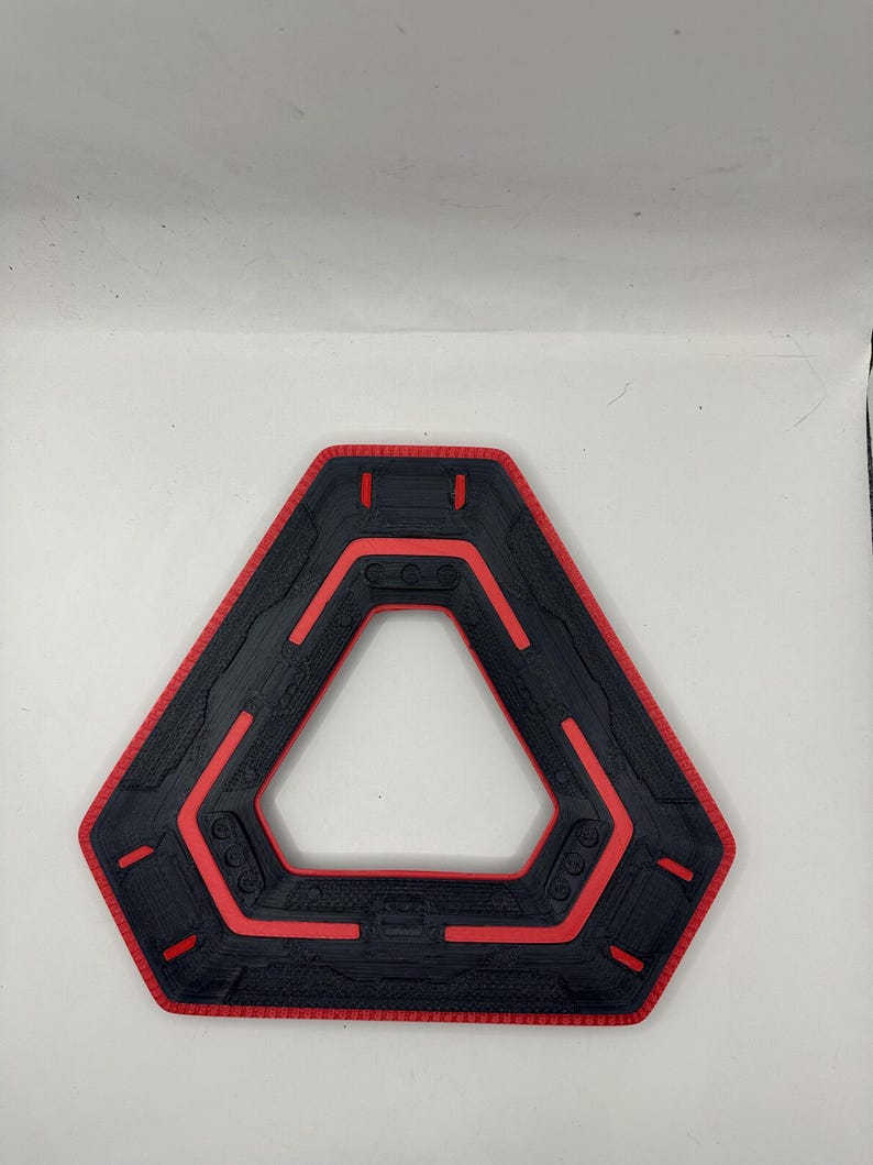 May include: A black and red geometric object with a triangular shape and a textured surface. The object has a red border and red accents within the black interior. The design features a series of parallel lines and rounded shapes.