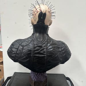 May include: A detailed bust of a character with a pale complexion, adorned with numerous metal pins embedded in the head. The figure wears a black, textured leather-like garment. The bust is mounted on a decorative base.