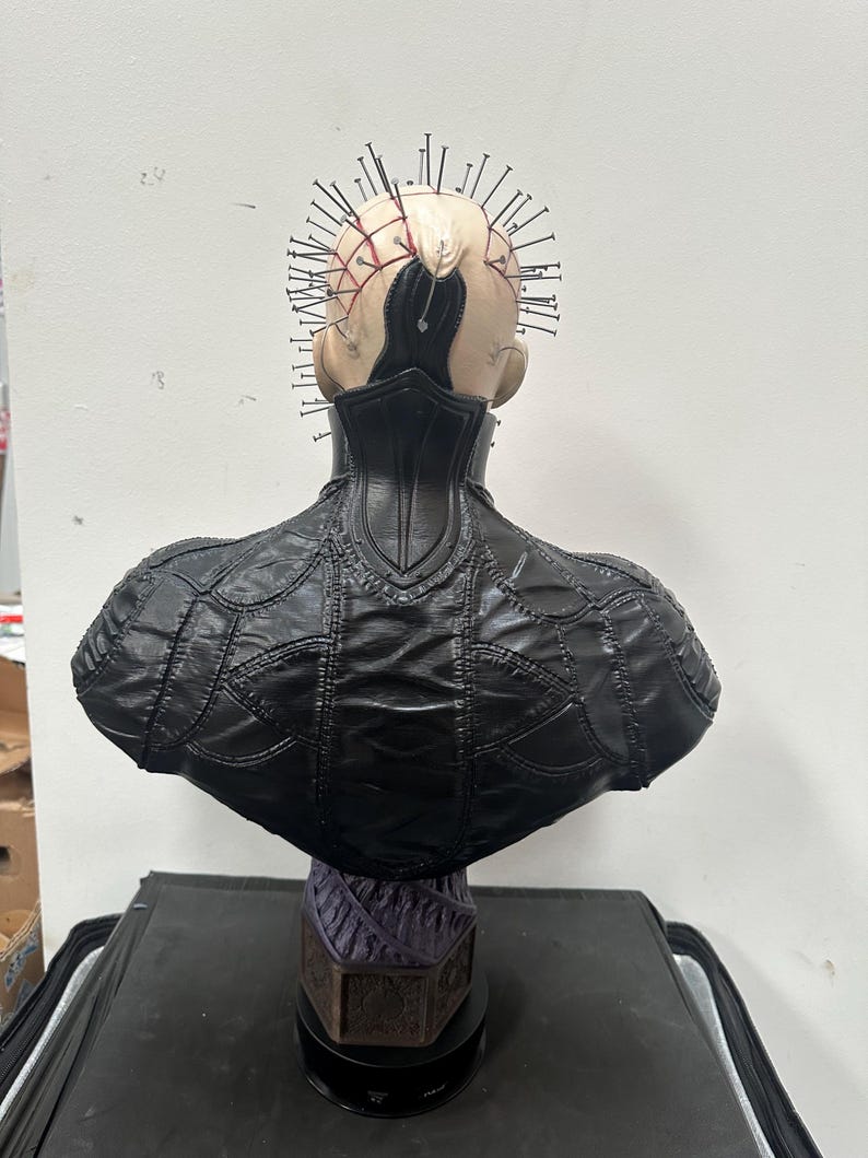 May include: A detailed bust of a character with a pale complexion, adorned with numerous metal pins embedded in the head. The figure wears a black, textured leather-like garment. The bust is mounted on a decorative base.