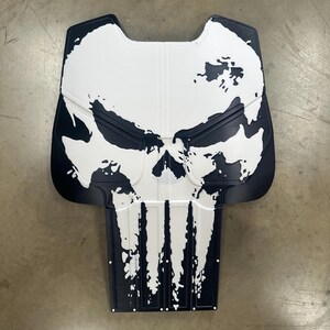 May include: A black and white graphic of a skull, resembling the Punisher logo. The design is on a shield-shaped object, with the skull's features prominently displayed. The background is a neutral gray.