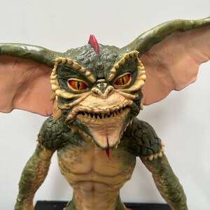 May include: A detailed figurine of a Gremlin creature. The figure has a green and tan color scheme with large ears, yellow eyes, and a red mohawk. The creature's mouth is open, revealing sharp teeth and a red tongue. The figure is posed with its arms at its sides.
