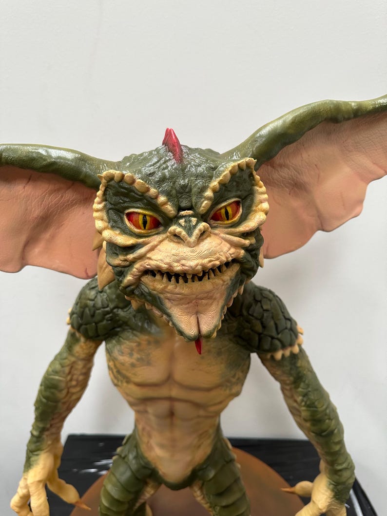 May include: A detailed figurine of a Gremlin creature. The figure has a green and tan color scheme with large ears, yellow eyes, and a red mohawk. The creature's mouth is open, revealing sharp teeth and a red tongue. The figure is posed with its arms at its sides.