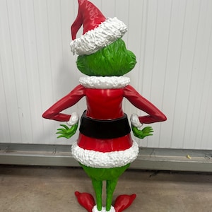 May include: A green figure with a red Santa hat and red outfit, trimmed with white faux fur. The figure has a black belt and is posed with arms out. The figure's feet are large and red, with white trim, and it is a Christmas decoration.