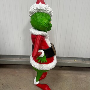 May include: A green character statue wearing a red Santa suit with white trim and a matching hat. The figure has a black belt and is posed in a side view, with a mischievous expression. A festive holiday decoration.