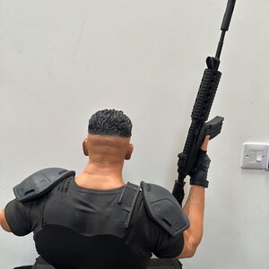 May include: A detailed bust of a figure in a black tactical shirt and shoulder pads, holding a black rifle. The figure's hair is neatly styled, and the overall design suggests a military or action theme.